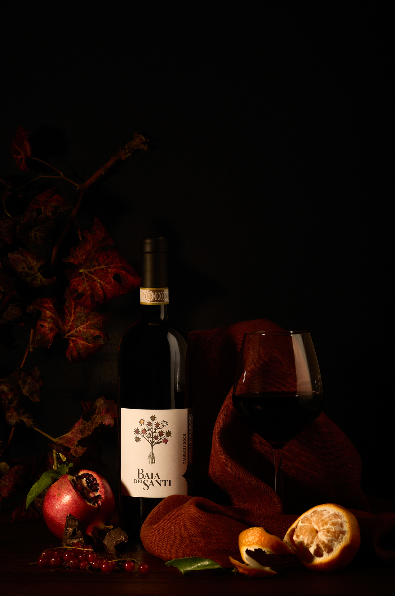 Bottiglia del vino Taurasi DOCG di Baia DeiSanti con calice, foglie, melograno, mandarino e ribes